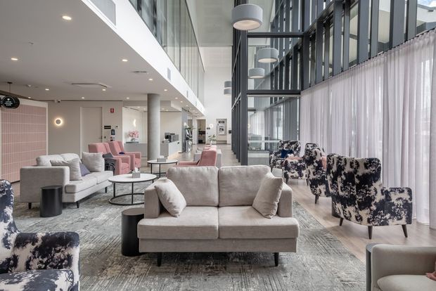 A bright, contemporary commercial lounge or reception area with multiple upholstered sofas and armchairs arranged on a large patterned rug. The space features a high glazed wall with sheer curtains, exposed dark window framing, cylindrical pendant lights, recessed ceiling lighting, concrete columns, and a pink tiled feature wall. The furniture uses soft neutral upholstery with blush-pink and bold black-and-white patterned accent chairs.