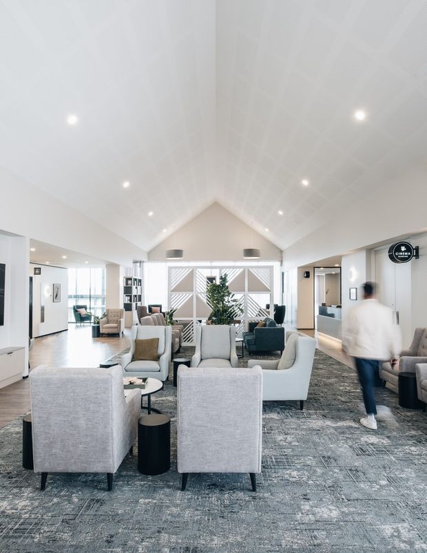 Bright, spacious communal lounge or waiting area with a dramatic white vaulted ceiling, extensive recessed lighting, pale walls, grey patterned carpet, and multiple upholstered lounge chairs arranged around small round tables. Glazed openings, abstract geometric screening, indoor planting, and a visible cinema sign suggest a contemporary cinema or hospitality foyer.