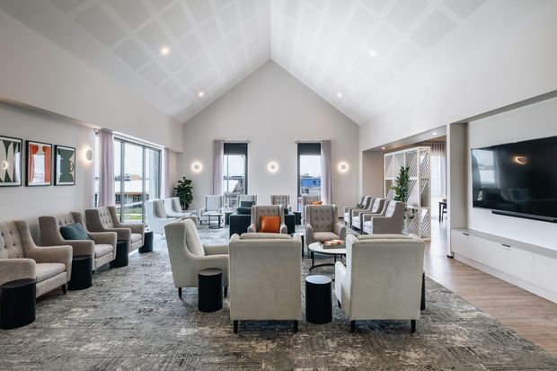 A spacious, bright contemporary living room arranged with multiple upholstered armchairs around small round tables on a large patterned rug. The room has a dramatic vaulted ceiling with a white geometric grid pattern, tall windows and glazed doors, pale neutral walls, integrated white media cabinetry with a large television, contemporary wall sconces, and a timber floor. A partial dining or adjacent seating area is visible at the rear.