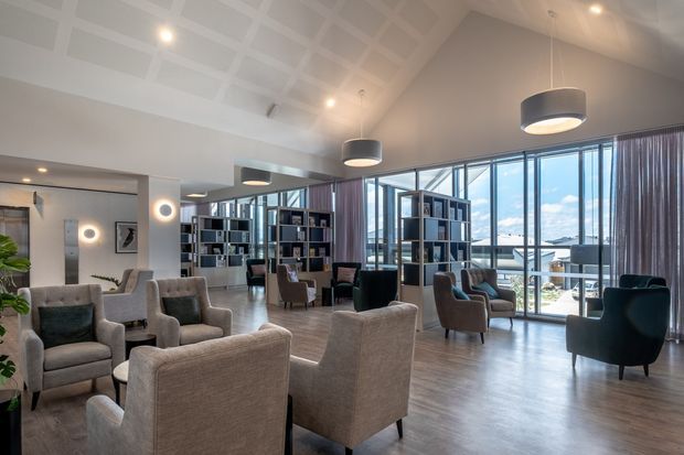 A spacious contemporary lounge or lobby with grouped upholstered armchairs, dark shelving and room dividers, large floor-to-ceiling windows, and pendant lighting. The room has a high coffered ceiling, warm grey timber-look flooring, and views toward an outdoor terrace or urban setting beyond the glazing.