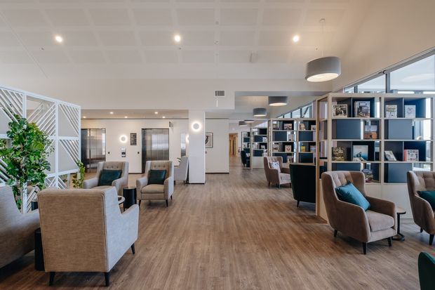 A bright, spacious contemporary lounge or shared communal area with warm timber-look flooring, extensive open shelving, upholstered armchairs, pendant lights, and large windows. White partition screens, recessed ceiling panels, and integrated display shelving create an airy, refined interior suitable for a hospitality, workplace, or aged-care setting.