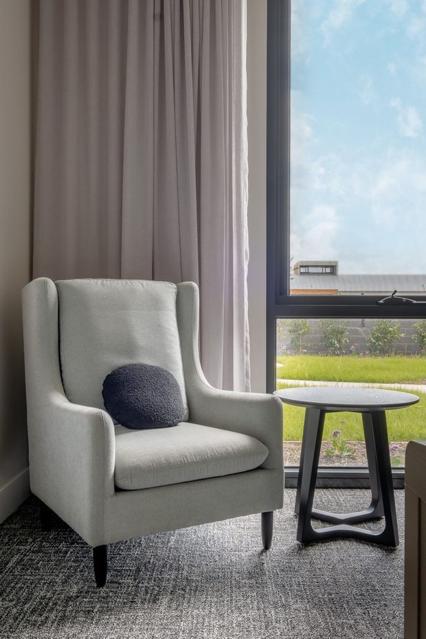 A calm, softly lit interior corner featuring a light upholstered wingback armchair with a dark round cushion, positioned beside a small dark side table. Floor-to-ceiling sheer curtains frame a large black-framed window looking onto a lawn and neighbouring building. A textured dark grey carpet covers the floor.