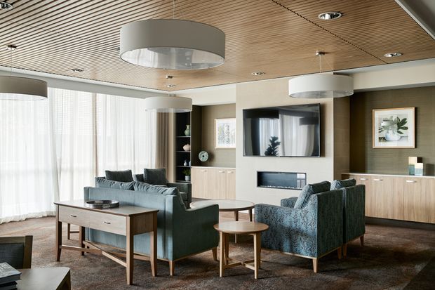 A refined contemporary living room with a dark upholstered sofa and armchairs arranged around nested timber coffee tables and a slim console table. The room features extensive sheer-curtained glazing, warm timber slatted ceiling, recessed downlights, large drum pendant lights, built-in timber cabinetry, a wall-mounted television, fireplace, and neutral textured finishes.