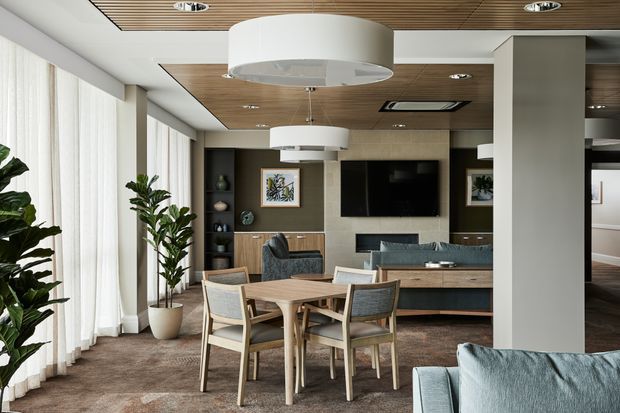 Contemporary open-plan living and dining area with a round timber dining table and upholstered chairs in the foreground, a blue-grey sofa, built-in timber cabinetry, a wall-mounted television, and decorative shelving. The room has full-height sheer curtains, a warm timber slatted ceiling with recessed lighting, pale textured wall surfaces, large white cylindrical pendant lights, and indoor plants.
