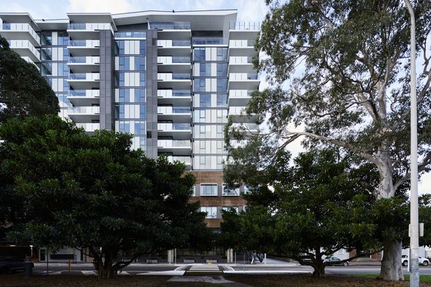 Exterior photograph of a contemporary multi-storey apartment building viewed from a landscaped street frontage. The facade features white horizontal slab forms, extensive floor-to-ceiling glazing, projecting balconies, dark grey vertical cladding, and lower-level brown brickwork, partially screened by mature trees.
