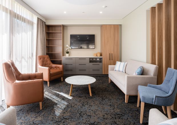 Bright, refined living room with a large window dressed in sheer curtains and drapes, a warm timber built-in entertainment wall with open shelving and concealed storage, a grey media console, and a wall-mounted television. The room is furnished with leather and upholstered armchairs, a pale sofa, a round marble-look coffee table, and a dark patterned rug.