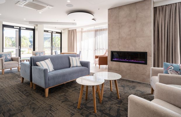 Bright, contemporary living room with a central fireplace feature wall, blue upholstered sofas and armchairs, nested round coffee tables, full-height curtains, large glazed doors and windows, and a patterned dark carpet. The space appears to be a refined shared lounge or residential sitting area with abundant natural light.