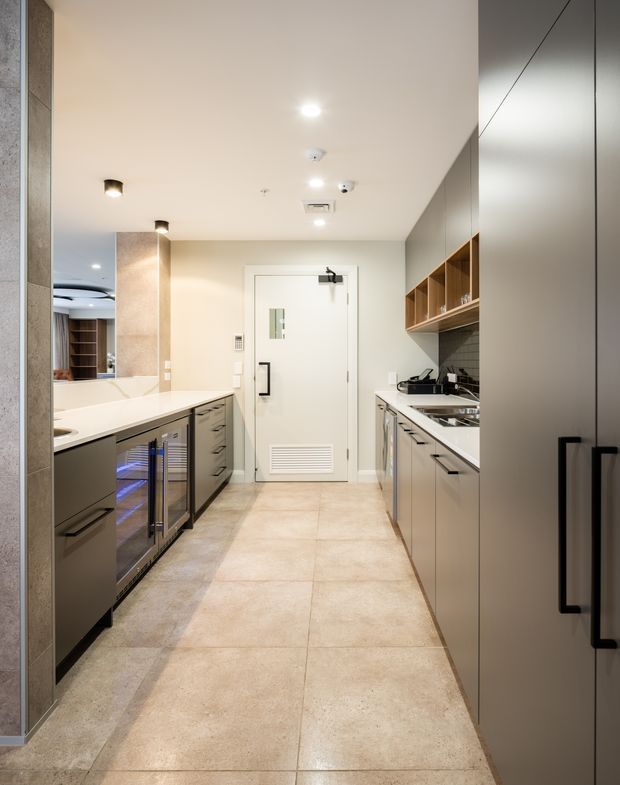 A compact, galley-style commercial kitchen or catering preparation area with parallel runs of grey fixed cabinetry, pale benchtops, integrated appliances, open timber shelving, a tiled splashback, and large beige floor tiles. The space has a clean contemporary fit-out, recessed ceiling lighting, stainless-steel equipment, and a rear service door.