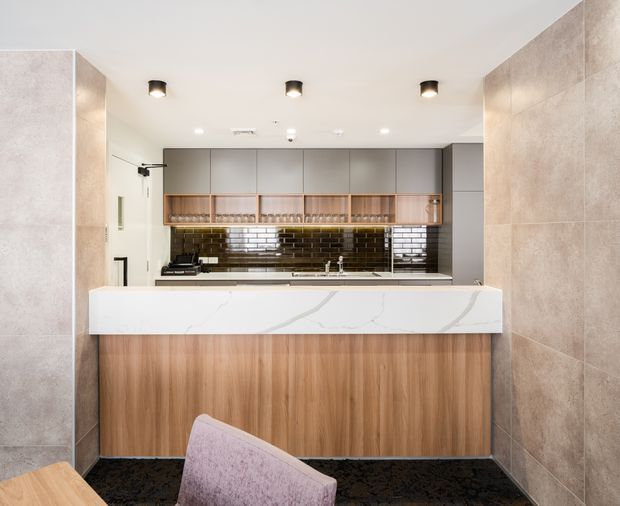 A contemporary commercial-style kitchenette or bar area viewed from the adjoining space. The room features a large white veined stone island benchtop with timber veneer cabinetry, grey overhead cabinets, open timber shelving with glassware, a glossy dark green subway-tile splashback, pale large-format tiled walls, recessed ceiling lights, and dark flooring. A partial upholstered chair is visible in the foreground.