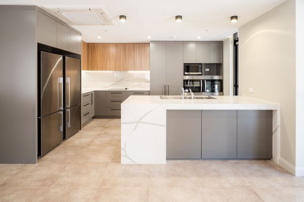 A contemporary kitchen with a large waterfall-edge island, integrated stainless-steel appliances, matte grey cabinetry, warm timber wall cabinetry, and a white veined stone benchtop and splashback. Large beige-toned floor tiles and recessed ceiling lighting create a clean, refined, neutral interior.