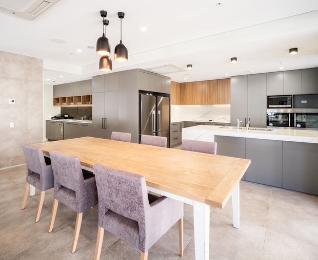 A bright, open-plan contemporary kitchen and dining area. The kitchen features extensive matte grey cabinetry, integrated stainless-steel appliances, a white island benchtop, warm timber cabinetry accents, and a large light timber dining table with upholstered dining chairs. Three dark pendant lights hang above the table, while large-format light neutral floor tiles and white ceilings create an airy, refined interior.
