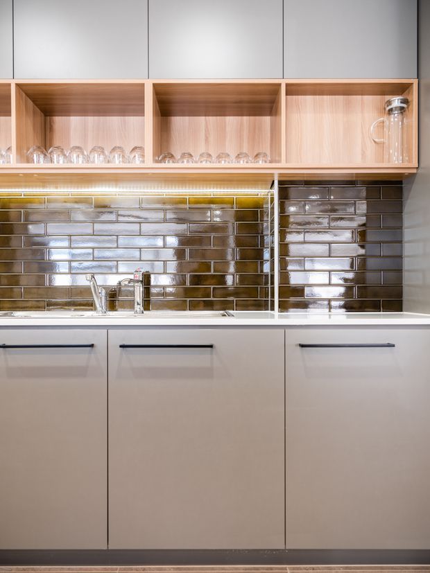 Close-up view of a contemporary kitchen run with matte pale cabinetry, dark linear handles, a white benchtop, glossy brown-grey subway tile splashback, stainless steel tapware, and open timber shelving displaying glassware. Integrated under-shelf lighting illuminates the work area.