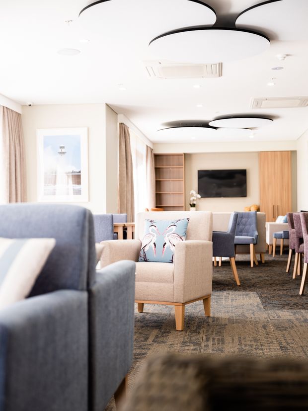 Bright, contemporary lounge or waiting area furnished with upholstered armchairs and sofas in blue, beige, and muted mauve tones. The room features patterned carpet, full-height curtains, light timber built-in cabinetry and shelving, a wall-mounted television, framed artwork, and distinctive overlapping circular ceiling light fixtures with dark trim.