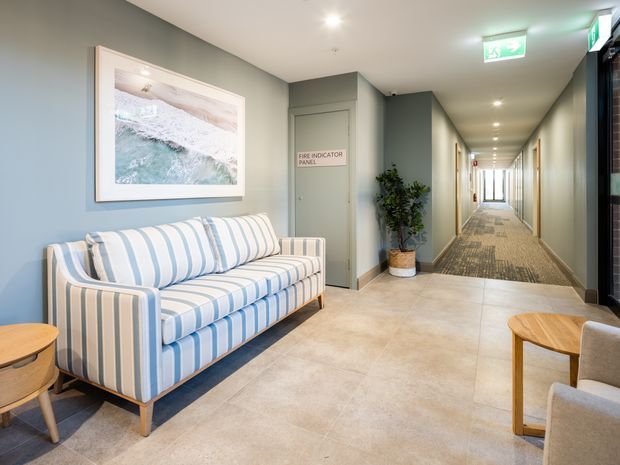 A bright, contemporary hotel or commercial lobby lounge with a striped blue-and-white sofa, small timber side tables, upholstered chairs, pale tiled flooring, muted grey-green walls, and coastal artwork. A carpeted corridor with recessed ceiling lighting and exit signage extends into the background.