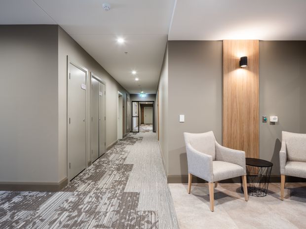 A modern hotel or commercial corridor with multiple doors, patterned carpet, recessed ceiling lights, and a small waiting lounge. The lounge features upholstered armchairs, a round black side table, and a vertical timber feature panel with integrated wall lighting.