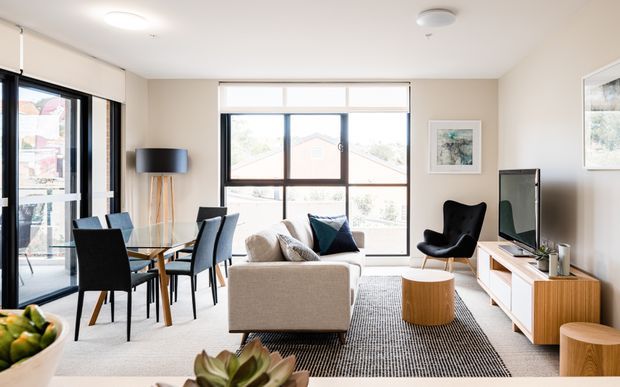 Bright contemporary open-plan living and dining area with large windows and glazed doors, a neutral sofa, black accent armchair, glass dining table with upholstered chairs, timber media console, and layered rugs. The room has abundant natural light and views to the outdoors.