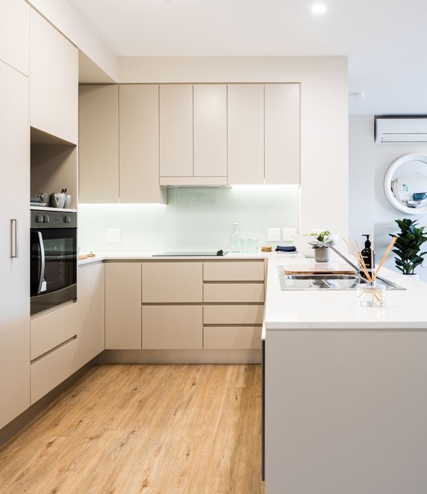 Bright, compact contemporary kitchen with full-height matte beige cabinetry, integrated black oven, pale stone or engineered-stone benchtops, a white glass backsplash, and warm timber-look flooring. A peninsula with a built-in cooktop occupies the foreground, while a round wall mirror and wall-mounted air conditioner are visible to the right. The space is clean, streamlined, and softly lit.