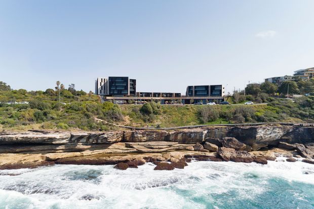 Wide exterior coastal view of a contemporary multi-storey building complex set on a landscaped rocky cliff above the ocean. The development features dark glazed volumes, horizontal connecting structures, and a long low-profile form integrated into the elevated site. Waves, exposed layered rock, coastal vegetation, and neighbouring buildings are visible.