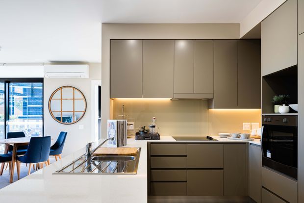 Contemporary open-plan kitchen with a large light stone island and integrated sink, matte taupe-brown full-height cabinetry, warm under-cabinet lighting, and built-in black appliances. A dining area with blue upholstered chairs is visible to the left, along with a large window, wall-mounted air conditioner, and round wall mirror.