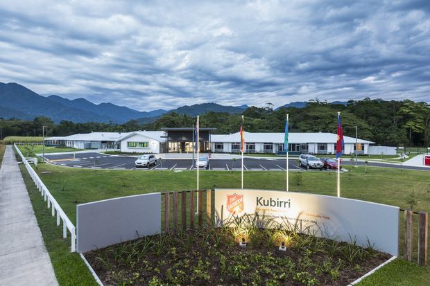 A low, modern institutional or commercial building viewed from the landscaped entrance. The site includes a curved Kubirri-branded sign, planted garden bed, flagpoles, marked parking bays, pedestrian paths, and a surrounding green landscape with forested mountains in the background. The building has a long horizontal form, light-coloured walls, dark roof sections, and extensive glazing beneath an overcast sky.