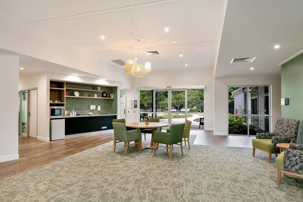 Bright, open-plan communal living and dining area with a compact integrated kitchenette along the left wall. The space features light timber-look flooring around the kitchen, pale carpet, white ceilings and walls, green upholstered dining and lounge chairs, patterned carpet, pendant lights, and extensive glazing opening toward a landscaped exterior.