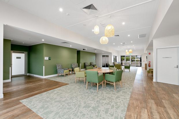A bright communal lounge or waiting area with green accent walls, pale patterned carpet, warm timber-look flooring, and clusters of upholstered armchairs and round tables. Woven pendant lights hang above the seating, while large glazed doors at the rear provide daylight. The space has a clean, contemporary commercial or aged-care interior with accessible circulation and multiple doors.