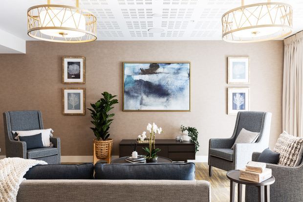 A refined living room with two grey upholstered lounge chairs, a matching sofa, a dark timber sideboard, framed artwork, indoor plants, and a round side table. The room has warm taupe walls, a patterned acoustic ceiling, pale curtains, and oversized gold-toned circular pendant lights.