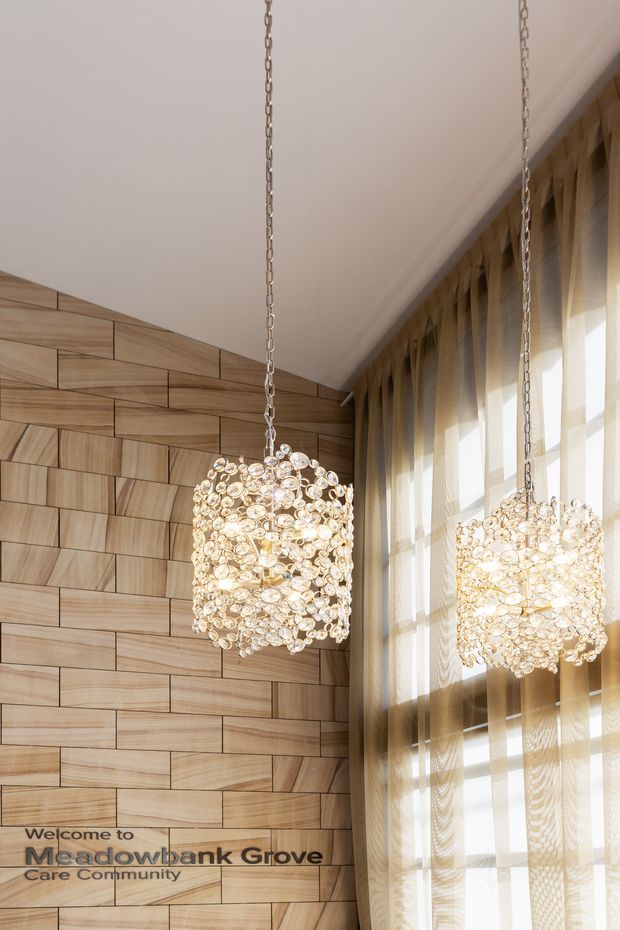 Interior architectural detail of a care community reception or common area, featuring two suspended decorative pendant lights made from clustered translucent discs or crystals. The space has a tall angled ceiling, a sculptural light timber feature wall with geometric plank arrangement, and full-height windows dressed with sheer curtains. Text on the wall reads “Welcome to Meadowbank Grove Care Community.”