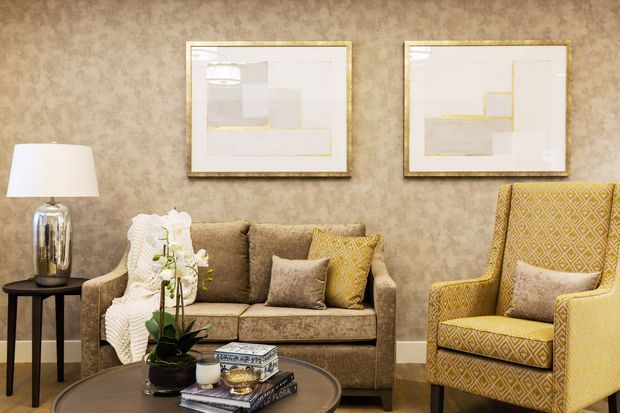 A refined living room vignette featuring a patterned upholstered sofa and armchair, a dark round coffee table with books and an orchid, a side table with a table lamp, framed abstract artwork, and textured taupe wall covering. The palette is warm neutrals with mustard-yellow accents and metallic details.