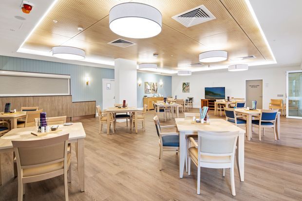 Bright contemporary dining and communal seating area with numerous light timber tables and chairs, blue upholstered accents, pale wood-look flooring, a slatted timber ceiling with concealed perimeter lighting, and large circular pendant lights. The space includes a built-in service or counter area along the left wall and appears to be a commercial hospitality or care setting.