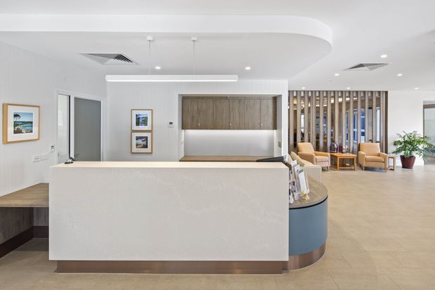 Bright contemporary reception area with a large white curved-front reception desk, integrated timber feature cabinetry, pale tiled flooring, white walls and ceiling, pendant and recessed lighting, and a timber slatted screen separating a small seating area. The space has a clean, refined commercial interior fit-out with blue and timber accents and indoor planting.