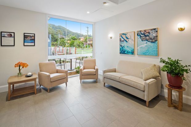 Bright, neatly furnished living room with a cream sofa, two beige armchairs, a timber side table, indoor plant, wall art, and subtle wall sconces. Large glazing opens the room visually to a small balcony or terrace and a leafy hillside beyond. The room has pale tiled flooring, light neutral walls, and a clean contemporary feel.
