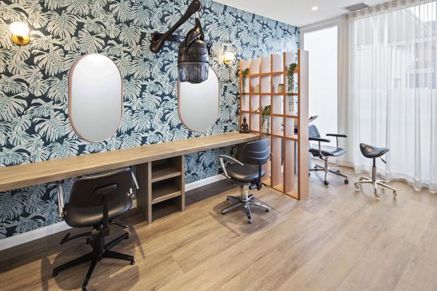 Bright contemporary salon or beauty studio with multiple styling stations, timber-look flooring, a long timber workbench with integrated open shelving, black salon chairs, oval mirrors, botanical patterned wallpaper, a timber open divider, and sheer floor-to-ceiling curtains. Decorative pendant lighting and wall sconces add a refined, playful character.