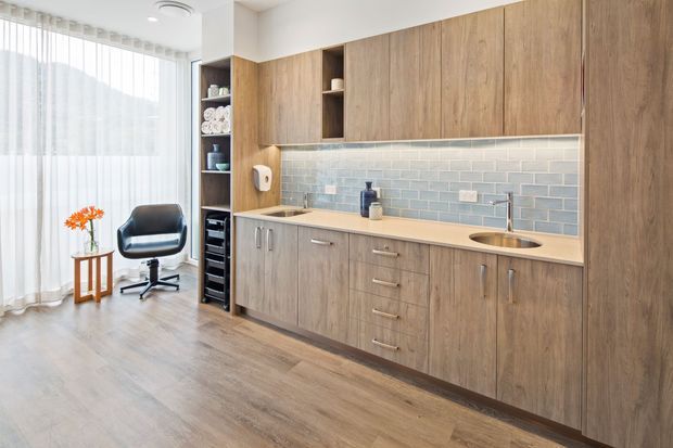 Bright, compact contemporary kitchenette or bar area with extensive timber-look cabinetry, a pale engineered-stone benchtop, blue-grey subway-tile splashback, integrated sink and open shelving. A small desk or work nook with a black swivel chair sits beside full-height sheer curtains, and the space has warm wood-look flooring and abundant natural light.
