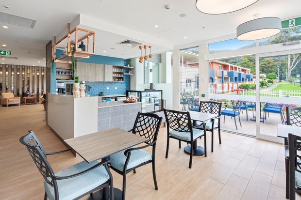 Bright contemporary café or hospitality dining area with a compact service bar and open kitchen zone. The space features pale tiled flooring, timber-look flooring, white cabinetry, a blue tiled counter backsplash, black metal-framed dining chairs and tables, pendant lighting, and full-height glazing opening to an outdoor terrace and landscaped setting.