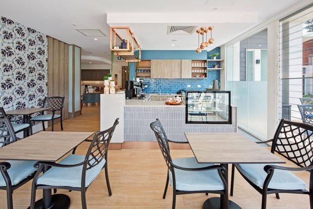 Bright contemporary café or hospitality interior with multiple dining tables and black-framed chairs, a compact service counter and display case, turquoise blue tiled backsplash, timber cabinetry, patterned feature wall, and large glazed windows. The space has a light, airy palette with timber flooring and colourful pendant lighting.