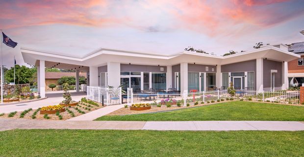 Single-storey contemporary residential building with a broad low-pitched roof, pale rendered walls, extensive glazing, covered outdoor areas, and a landscaped front garden. The accessible frontage, gentle paths, fencing, and level site suggest an age-friendly or assisted-living setting. A flagpole and neighbouring homes are visible in the background beneath a colourful sunset sky.