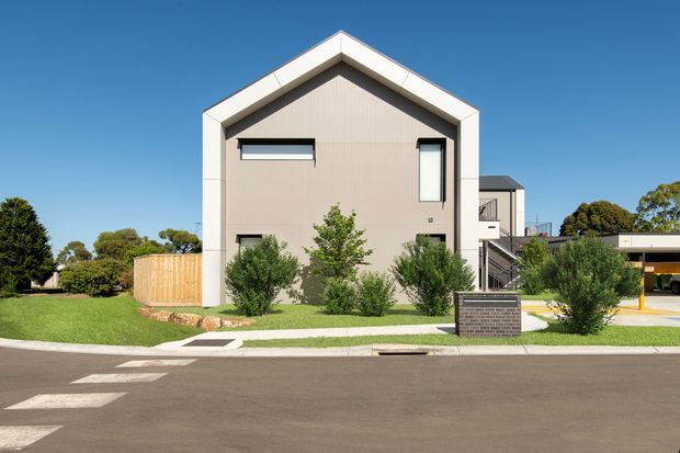 Street-facing view of a contemporary multi-storey residential building with a prominent gable-front form, pale rendered facade, white fascia and trim, dark window frames, landscaped front garden, pedestrian path, timber fence, and an adjacent carport structure. The image is captured from the street in clear daylight.