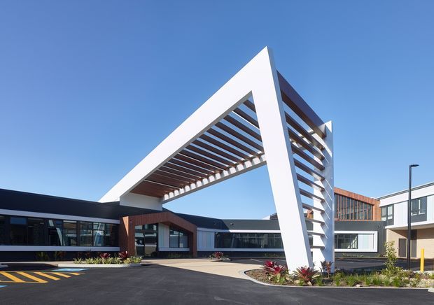 Exterior view of a contemporary low-rise commercial or institutional building complex, featuring a dramatic white triangular entry canopy with timber-like horizontal sun-shading slats. The building has dark and white cladding, extensive glazing, brick-toned sections, landscaped planting beds, and a broad paved forecourt under a clear blue sky.