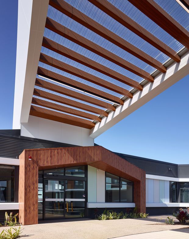 Exterior view of a contemporary low-rise building entrance beneath a large timber pergola. The facade combines white rendered surfaces, dark horizontal cladding, extensive dark-framed glazing, and a projecting timber-clad entry portal. Strong linear roof beams with translucent panels create a shaded covered approach, while low landscaping and paving sit in front of the entrance under a clear blue sky.