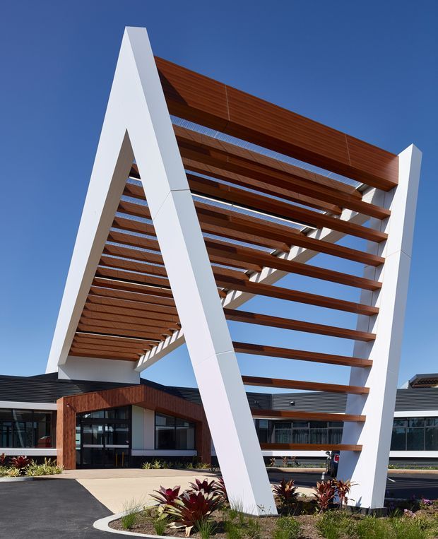 Exterior architectural photograph of a contemporary low-rise building featuring a dramatic white A-frame pergola or entry canopy with extensive exposed timber slats. Dark horizontal cladding, glazed entrances, landscaped planting, and paved driveways are visible beneath a clear blue sky.