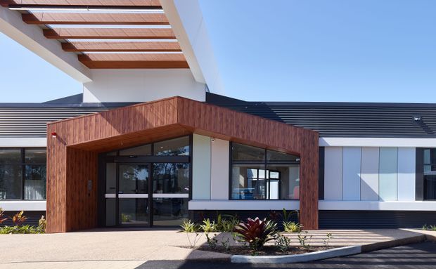 Exterior view of a contemporary, low-rise building entrance. The facade combines dark horizontal cladding, white rendered walls, extensive dark-framed glazing, and a prominent warm timber-clad entry canopy. A pergola with exposed timber beams and translucent roofing projects above the entrance, while a simple paved forecourt and planted garden bed sit in front.