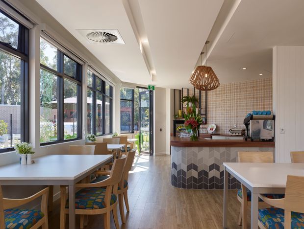 Bright contemporary café-style dining and service area with large black-framed windows, timber tables and chairs, a tiled service counter, patterned tiled backsplash, pendant lighting, and an espresso machine. The space opens to a landscaped exterior through extensive glazing and a glazed door.