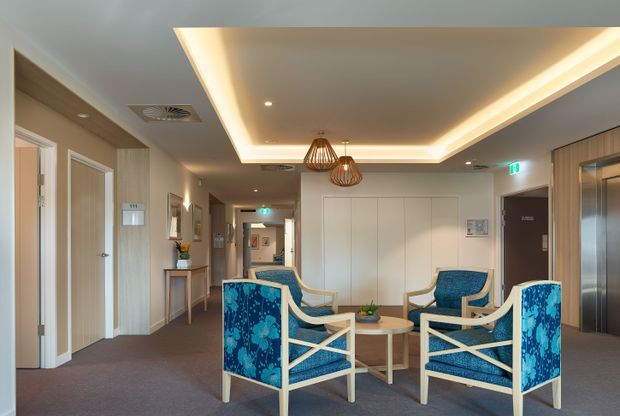 A bright, refined communal lounge or waiting area in a contemporary building, furnished with four blue patterned upholstered armchairs around small round tables. The space has warm beige and light timber wall finishes, carpeted flooring, recessed ceiling lighting with concealed LED cove illumination, decorative pendant lights, lift doors, and corridor or room entrances. The setting appears hospitality or healthcare-oriented, with a calm, welcoming atmosphere.