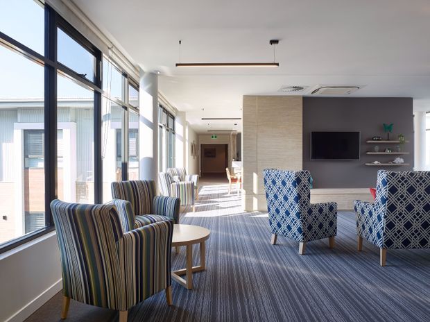 A bright contemporary communal lounge or waiting area with floor-to-ceiling windows along one side, striped upholstered armchairs, patterned blue accent chairs, small round tables, and a wall-mounted television with open shelving. A corridor extends beyond the seating area, and the space has carpeted flooring, neutral walls, and linear pendant lighting.