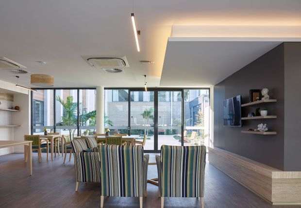 Bright contemporary open-plan living and dining area with floor-to-ceiling glazing opening to an outdoor courtyard or pool area. The room features striped upholstered dining chairs, a timber dining table, built-in shelving, a wall-mounted television, timber-look flooring, and a dark charcoal media wall with a projecting ceiling bulkhead and concealed LED lighting.