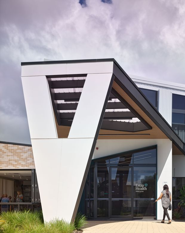 Exterior architectural photograph of a contemporary health facility entrance. The building features a bold white angular portal with dark trim, a dramatic sloping structural frame, timber-look soffit lining, extensive dark-framed glazing, and corrugated metal wall cladding. Paved entry landscaping and signage identify the entrance as Estia Health.