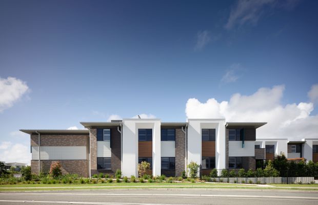 Street-facing view of a contemporary multi-unit residential building, featuring a long low-profile form, flat projecting rooflines, white rendered vertical volumes, dark grey and brown brickwork, broad windows, balconies, landscaped frontage, and a black metal fence. The image is taken from across the road under a bright blue sky.