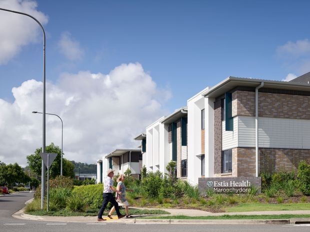 Exterior streetscape view of a contemporary low-rise healthcare building identified by signage as Estia Health Maroochydore. The building has stepped gabled roof forms, projecting white vertical frames, pale brick and horizontal white cladding, dark window frames, landscaped planting, and a pedestrian footpath beside the road.