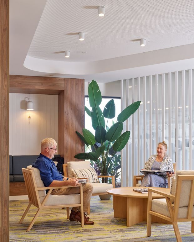Bright, contemporary lounge or waiting area with timber-framed upholstered armchairs arranged around a rounded timber coffee table. A built-in timber-lined alcove with a dark bench seat and wall sconces sits to the left, while white vertical screening and glazing define the background. A large indoor tropical plant adds greenery, and the patterned carpet introduces muted yellow, blue, and grey tones. The space has a clean, refined commercial or hospitality character with a curved, textured white ceiling and recessed spotlights.